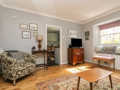 A living room with a floral armchair a wooden cabinet with a television a window seat with cushions and a wooden table at Primrose Cottage in Penryn