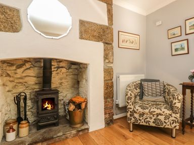 A stone fireplace with a wood burning stove next to a floral armchair and framed pictures on the wall at Primrose Cottage in Penryn