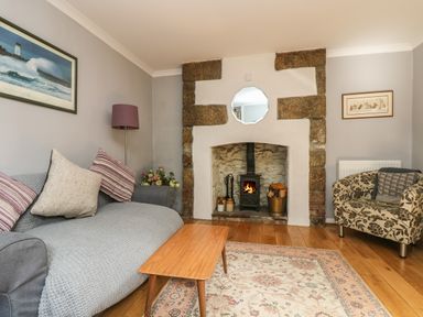 A living room with a gray sofa with cushions a wooden coffee table a patterned armchair and a fireplace with stones at Primrose Cottage in Penryn