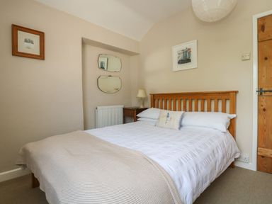A bedroom with a wooden bed frame white bedding a bedside table with a lamp and wall art at Primrose Cottage in Penryn