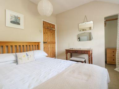 A bedroom with a wooden bed frame white bedding a wooden door a wooden desk with a stool two mirrors on the wall and a chest of drawers at Primrose Cottage in Penryn