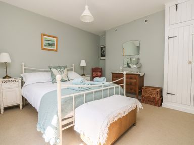 A bedroom with a white metal bed frame bedside tables with lamps a wooden chest of drawers a wicker storage basket and a white wooden wardrobe at Primrose Cottage in Penryn