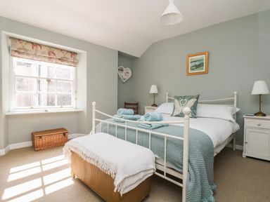 A bedroom with a white metal bed frame, two bedside tables with lamps, a window with floral blinds and a wicker chest at Primrose Cottage in Penryn