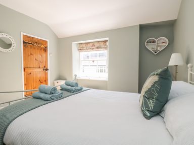 A bedroom with a bed a wooden door a window heart shaped wall shelf and a bedside lamp at Primrose Cottage in Penryn