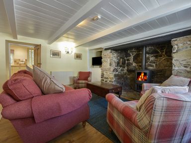 A living room with sofas and armchairs around a wood burning stove set in a stone fireplace at April Cottage in Coverack