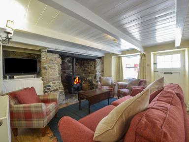 A living room with a stone fireplace wood stove red and plaid armchairs a red sofa a wooden coffee table a television on a stand and a door with windows at April Cottage in Coverack