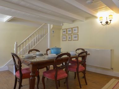 A dining room with a wooden table and six red cushioned chairs near a staircase at April Cottage in Coverack