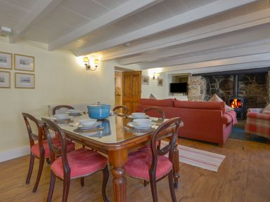 A dining area with a wooden table set with bowls and cutlery and a blue pot next to a living room with a red sofa and a fireplace at April Cottage in Coverack