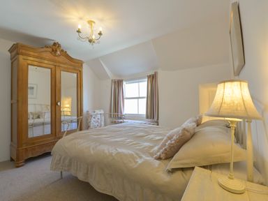 A bedroom with a bed a wooden wardrobe with mirrors a window with striped curtains and a lamp on a bedside table at April Cottage in Coverack