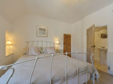 A bedroom with a metal bed frame, pillows, two bedside tables with lamps, wooden doors and an en suite bathroom at April Cottage in Coverack