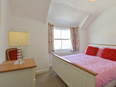 A bedroom with a white bed, red pillows, a bedside table with a lamp, and floral curtains at April Cottage in Coverack