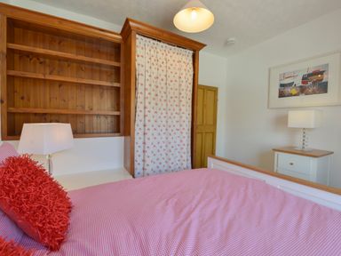 A bedroom with a bed covered with red and white bedding next to a wooden bookshelf and a curtain covering a wardrobe at April Cottage in Coverack