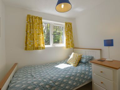A bedroom with a bed near a window with yellow curtains and a bedside table with a blue lamp at April Cottage in Coverack