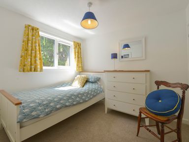 A bedroom with a single bed next to a window with yellow curtains a white dresser with a blue lamp and a wooden chair with a blue round cushion at April Cottage in Coverack
