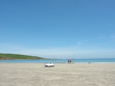 A sandy beach with a small inflatable boat and two people near the water with boats and a green hill in the background at April Cottage in Coverack