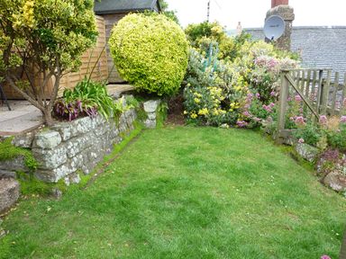 A garden with a stone retaining wall green lawn shrubs flowers and a wooden gate at April Cottage Coverack