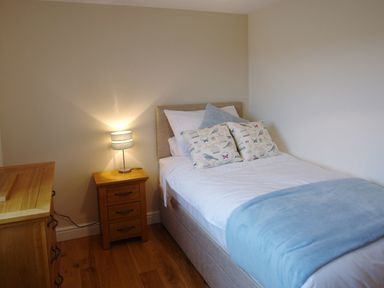 A bedroom with a single bed covered with white bedding and blue throw next to a wooden bedside table with a lamp at The Hayloft in Breage