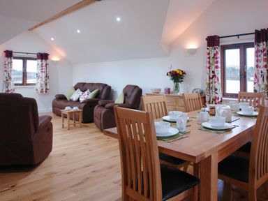A dining table set with white dishes and cups next to a seating area with brown leather sofas and floral curtains at The Hayloft in Breage