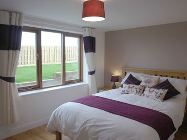 A bedroom with a bed featuring white and purple bedding next to a window with white curtains at The Hayloft in Breage