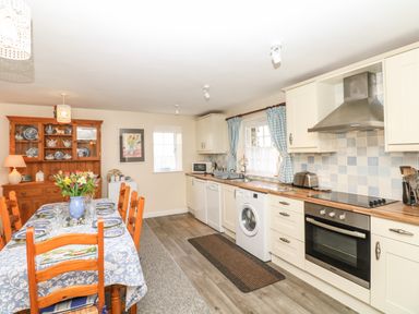 A kitchen with a dining table set with plates and flowers and cupboards with appliances at Badger Cottage in Mawgan-in-Meneage near Helston