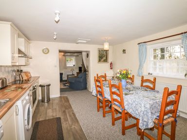 A kitchen area with wooden cabinets and appliances next to a dining table with six chairs and a flower vase at Badger Cottage in Mawgan-in-Meneage near Helston