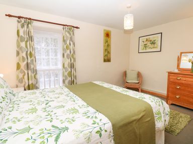 A bedroom with a double bed covered with floral bedding and a green throw near a window with leaf patterned curtains a wicker chair with cushions and a wooden dresser with mirror at Badger Cottage in Mawgan-in-Meneage near Helston