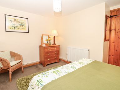 A bedroom with a bed green blanket wood chest of drawers lamp wicker chair and wall art at Badger Cottage in Mawgan-in-Meneage near Helston