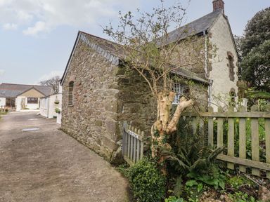 An exterior view of a stone and white cottage with a gray slate roof a wooden fence and small gate with plants and trees at Badger Cottage in Mawgan-in-Meneage near Helston