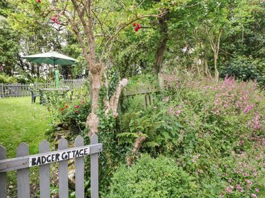 A garden with a wooden gate labeled Badger Cottage surrounded by flowers shrubs and trees with a table and umbrella in the background at Badger Cottage in Mawgan-in-Meneage near Helston