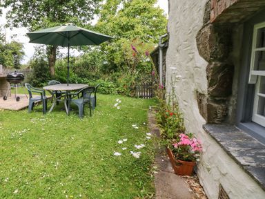 A garden with a round table and four plastic chairs under a green umbrella and a barbecue grill near the side of a white stone house at Badger Cottage in Mawgan-in-Meneage near Helston