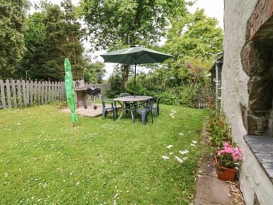 A garden with a round table and chairs under a green umbrella and a barbecue near a picket fence at Badger Cottage in Mawgan-in-Meneage near Helston