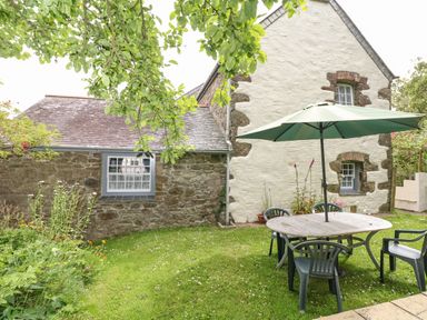 A garden with a round wooden table and four chairs under a green umbrella near a stone cottage with two windows at Badger Cottage in Mawgan-in-Meneage near Helston