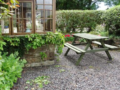 A garden area with a wooden picnic table gravel ground and plants near a window at Swift Cottage in Mawgan-in-Meneage near Helston