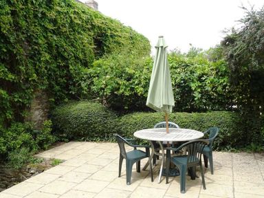 An outdoor patio with a round table and four plastic chairs and an umbrella surrounded by greenery and bushes at Swift Cottage in Mawgan-in-Meneage near Helston