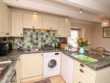 A kitchen with cream cabinets a washing machine a countertop with a vase of flowers and a dining table in the background at Swift Cottage in Mawgan-in-Meneage near Helston