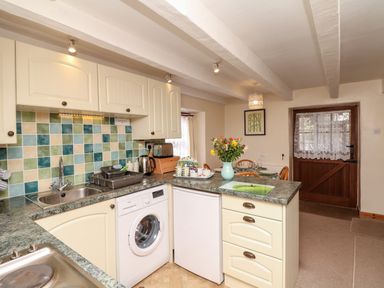 A kitchen with cream cabinets green tiled backsplash and dining area with flowers on table at Swift Cottage in Mawgan-in-Meneage near Helston