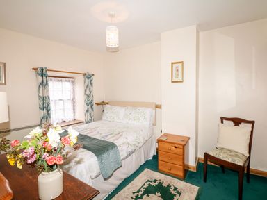 A bedroom with a double bed a wooden chest of drawers a wooden chair with cushion floral curtains and a vase of flowers on a wooden dresser at Swift Cottage in Mawgan-in-Meneage near Helston