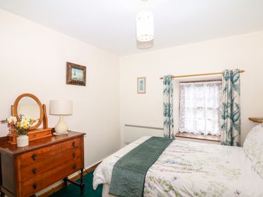 A bedroom with a wooden dresser with a mirror and flowers a bed with floral bedding and a window with curtains at Swift Cottage in Mawgan-in-Meneage near Helston