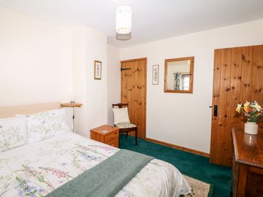 A bedroom with a double bed a chair wooden doors a mirror and a dresser with flowers at Swift Cottage in Mawgan-in-Meneage near Helston