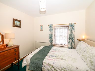 A bedroom with a bed with floral bedding green curtains and a wooden dresser with a lamp at Swift Cottage in Mawgan-in-Meneage near Helston