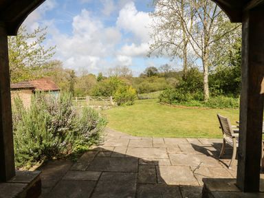 A patio with stone tiles and wooden chairs looking out to a garden with grass shrubs trees and a wooden gate at Marles Farmhouse in Salwayash
