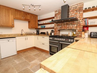 A kitchen with wooden upper cabinets white lower cabinets a black stove set in a brick alcove open shelves with dishes and cookbooks and a dishwasher at Marles Farmhouse in Salwayash