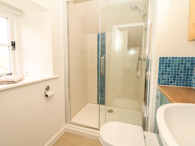 A bathroom with a glass shower enclosure, white toilet, sink, blue tiled backsplash, and a window with a towel on the sill at Marles Farmhouse in Salwayash
