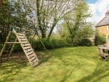 A garden with a wooden climbing frame and swing set alongside a wooden table and chairs outside a stone house at Marles Farmhouse in Salwayash