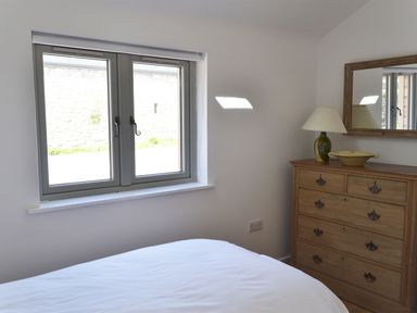 A bedroom with a bed under a double window and a wooden dresser with a lamp and bowl next to a mirror at The Studio Rinsey Cove near Praa Sands