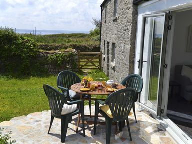 An outdoor patio with a round wooden table set for breakfast and four green plastic chairs next to a stone house at The Studio Rinsey Cove near Praa Sands