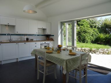 A kitchen with white cabinets and a dining table set with dishes next to a large open door showing a garden at The Studio in Rinsey Cove near Praa Sands
