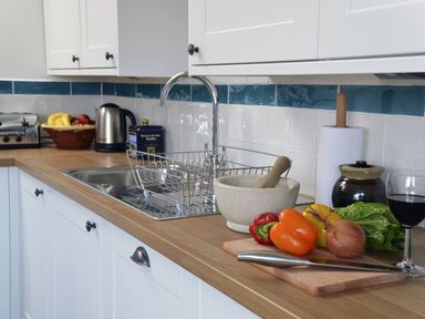 A kitchen counter with vegetables on a cutting board a knife a mortar and pestle a glass of wine and a sink at The Studio Rinsey Cove near Praa Sands