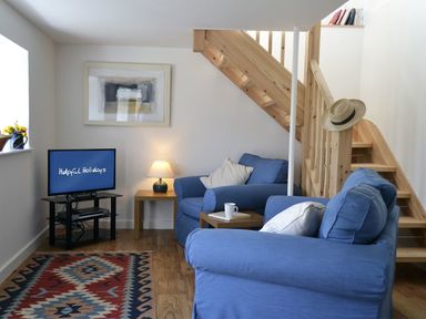 A living room with blue sofas a wooden staircase a television and a small wooden table with a lamp at The Studio in Rinsey Cove near Praa Sands