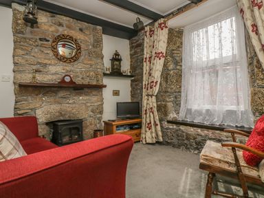 A sitting room with stone walls a fireplace a red sofa a wooden chair and a window with floral curtains at Mollys Cottage in St Just
